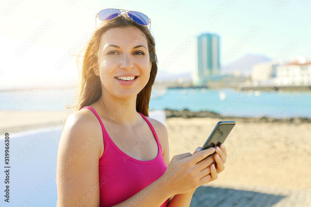 Beautiful trendy girl messaging with smart phone on seaside city looking at camera