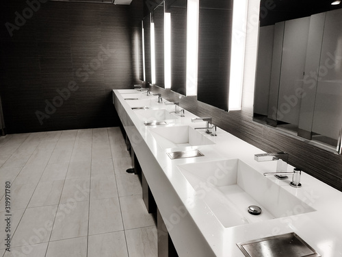 Public empty restroom with washstands, baby changer, and toillets in mirror. White sink row with mirrors and lights. Top horizontal view copyspace