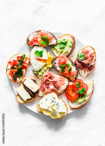 Variety of tapas sandwiches plate - sandwiches with prosciutto, avocado, salmon, egg, tomatoes, jamon, gorgonzola, brie, pear on a light background, top view. Delicious snack, appetizers. Copy space