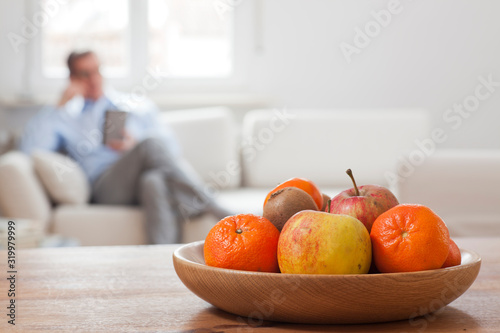 Fototapeta Naklejka Na Ścianę i Meble -  Mature man sitting with smartphone in a living room with fruit bowl in the foreground