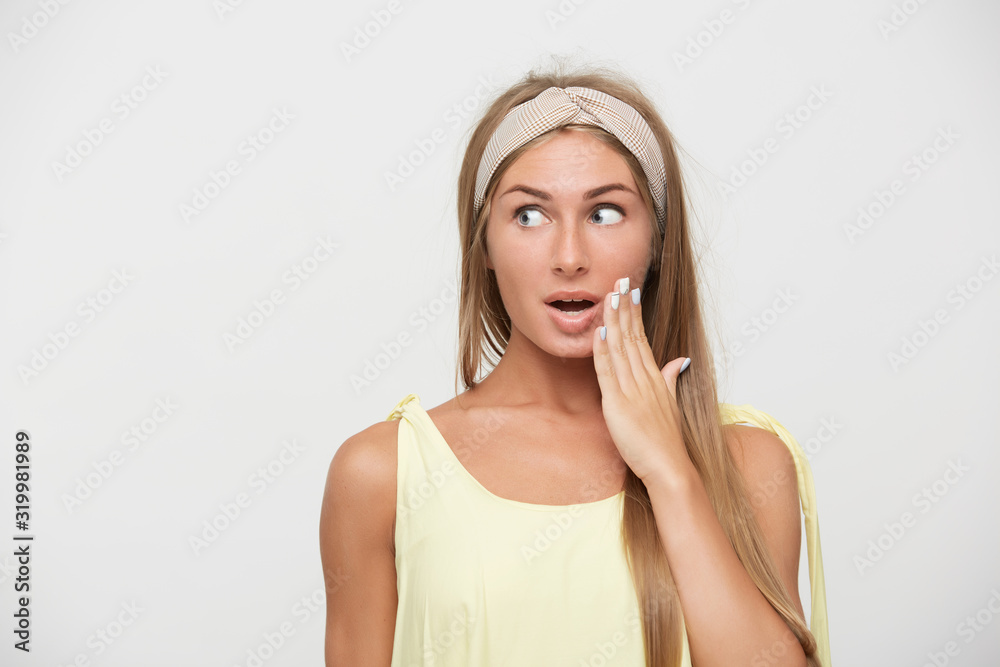 Agitated young pretty long haired blonde woman with casual hairstyle rounding her blue eyes while looking excitedly aside and keeping raised hand near her mouth, isolated over white background Agitated young pretty long haired blonde woman with casual hairstyle rounding her blue eyes while looking excitedly aside and keeping raised hand near her mouth, isolated over white background