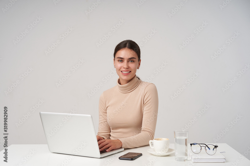 Indoor photo of cheerful young pretty brunette female showing her perfect white teeth while smiling positively at camera, typing text on keybord while posing over white background