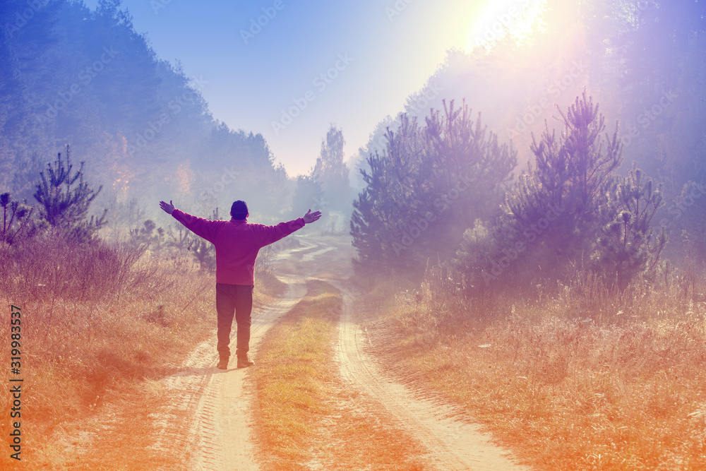 Happy man with hands in the air standing on a dirt country road in the forest early in the morning and looking at sunrise