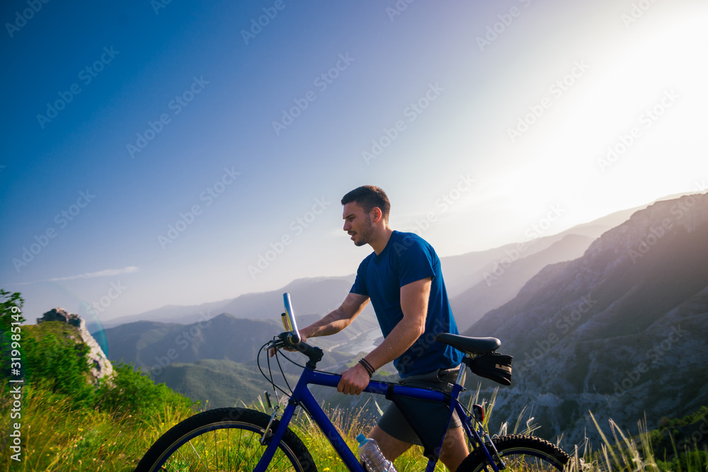 Fototapeta premium Perspective of a fit mountain biker pushing his bike uphill with amazing view on a forest, river and mountains in the background. Amazing green nature at sunset.