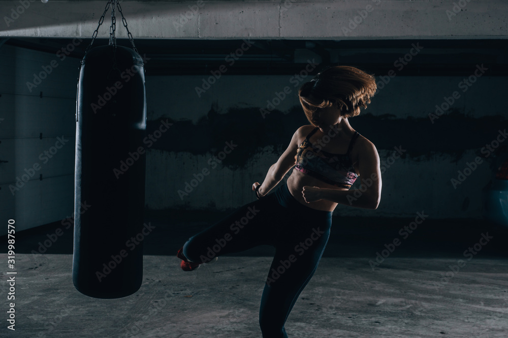 Young silhouette female practicing high kick with the boxing bag inside the garage.