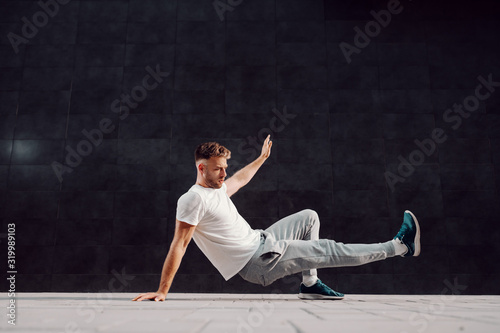 Attractive fit caucasian bearded muscular man in tracksuit doing leg kicks on the ground. In background is gray wall.