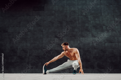 Full length of attractive Caucasian bearded blonde man stretching leg. In background is gray wall.