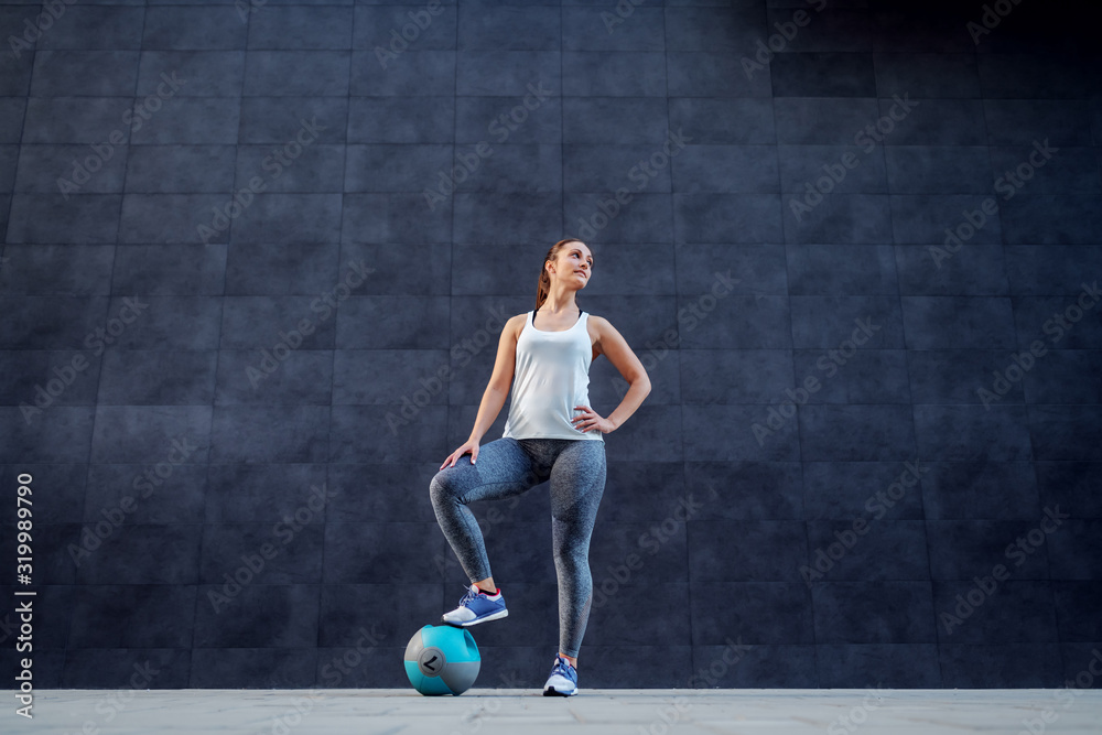 Fototapeta premium Attractive fit Caucasian brunette in sportswear holding foot on weight ball and posing outdoors. In background is dark wall.