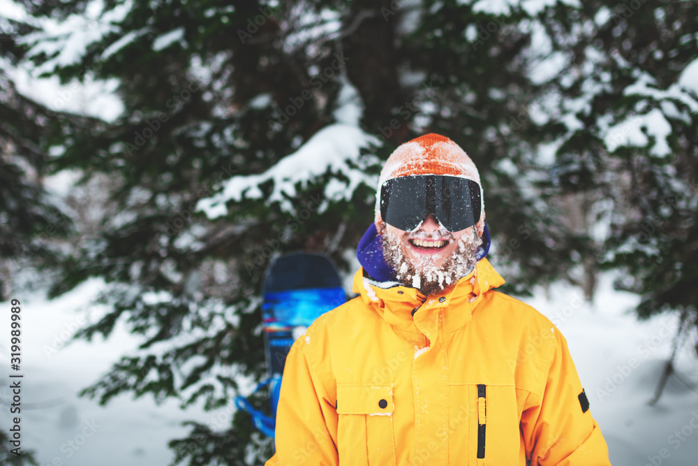 Close up portrait of bearded snowboarder or skier wearing black goggles and orange hat with snow on face standing in mountain forest and smiling. Happy people vacation.