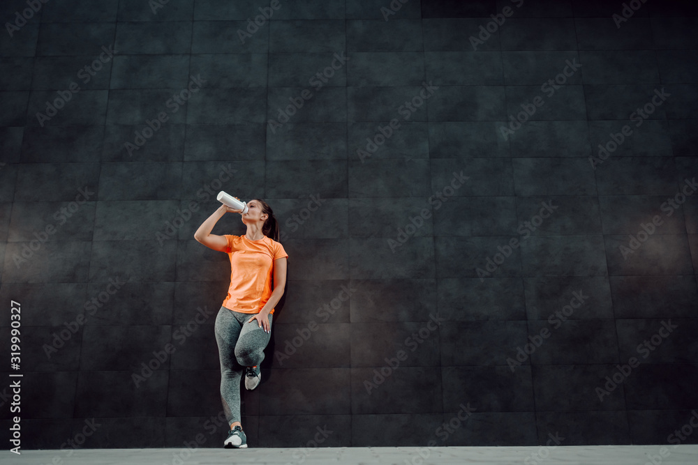 Full length of attractive caucasian brunette in sportswear and with ponytail leaning on wall and drinking fresh water.