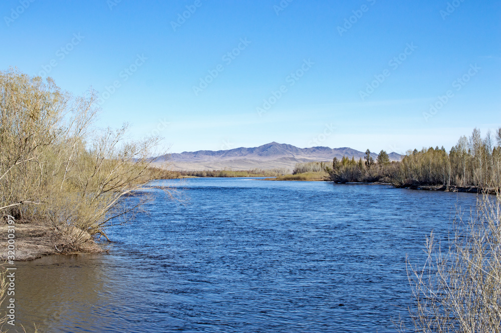 a large blue river spreads between the two banks against the backdrop of high mountains and blue sky