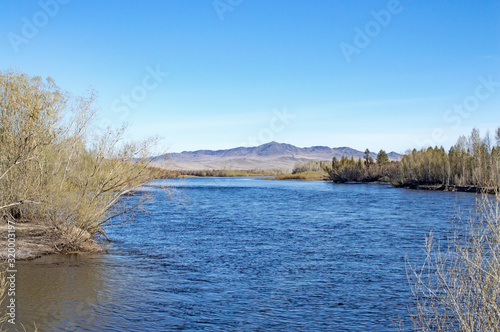 Fototapeta Naklejka Na Ścianę i Meble -  a large blue river spreads between the two banks against the backdrop of high mountains and blue sky