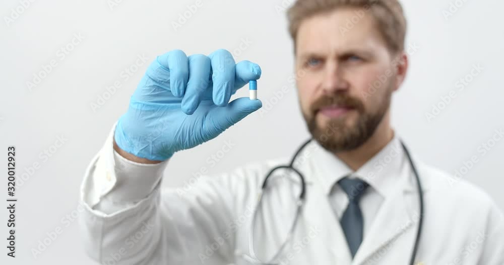 Close up of medical pill that holding bearded young doctor in white lab coat, rubber gloves in studio. Concept of presenting new medicines. Close up of medical pill that holding bearded young doctor in white lab coat, rubber gloves in studio. Concept of presenting new medicines.