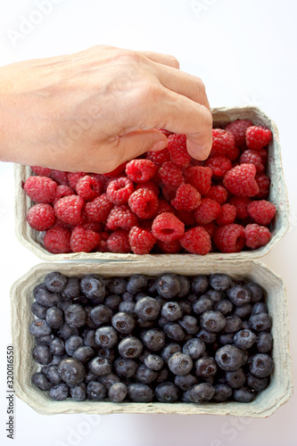 Photo of sweet raspberry and blueberry in packing containers. Woman's hand takes juicy berries from box. Red and blue fruit in package, closeup. Healthy, delicious dessert in packaging. Isolated.