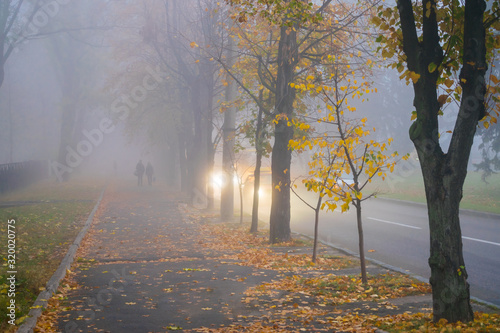 The sidewalk with a pair of walkers is in dense fog. Cars move along an autumn road in conditions of poor visibility. Bad weather on city street. Blurred fog effect.