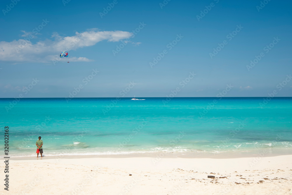 The turquoise sea and the white sand of the dolphin beach in Cancun Mexico, a man observes the horizon, in the background a parasailing with tourists in the blue sky