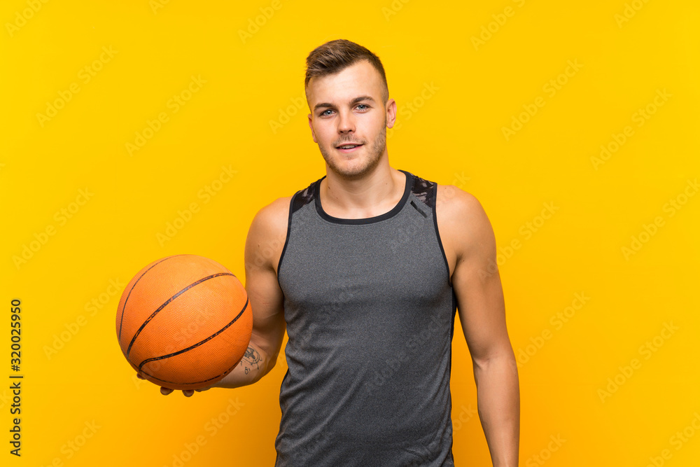 Young handsome blonde man holding a basket ball over isolated yellow background smiling a lot