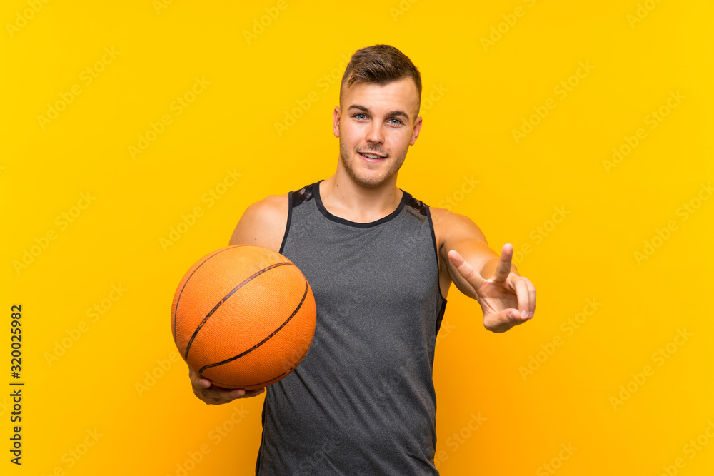 Young handsome blonde man holding a basket ball over isolated yellow background smiling and showing victory sign