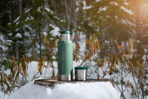 A green thermos stands on an old stump in the forest. The background of the forest is blurred.