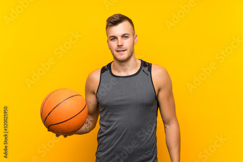 Young handsome blonde man holding a basket ball over isolated yellow background smiling a lot