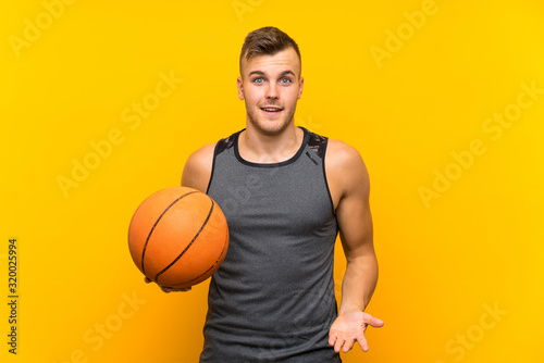 Young handsome blonde man holding a basket ball over isolated yellow background with shocked facial expression