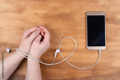 Hands tied with a cord of earbuds and a mobile phone on a wooden background (top view) as the mobile or cell phone addiction concept