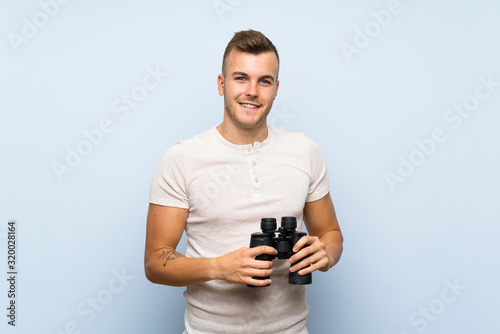 Young handsome blonde man over isolated blue background with black binoculars