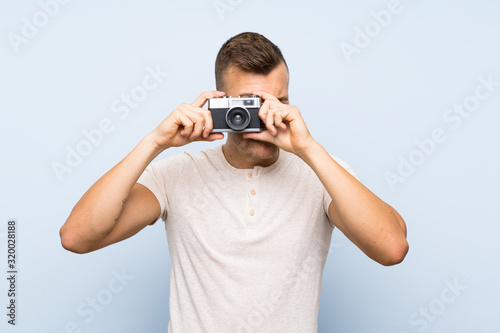 Young handsome blonde man over isolated blue background holding a camera