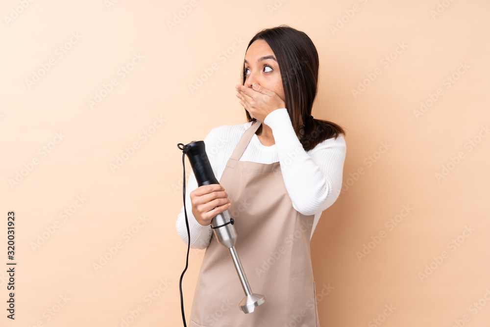 Young brunette girl using hand blender over isolated background covering mouth and looking to the side