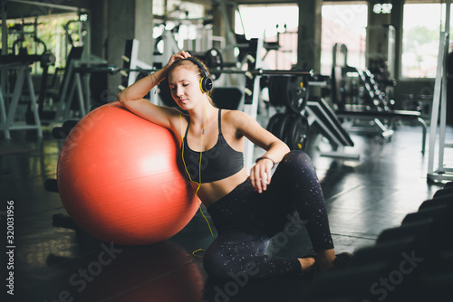 A beautiful woman wearing a sports shirt is sitting and relaxing by listening to music from headphones. With rubber ball, yoga or exercise ball
