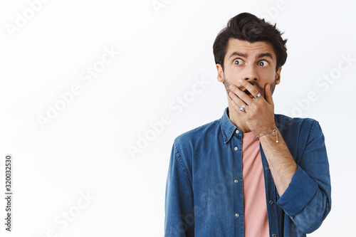 Shocked, scared handsome bearded male in shirt over pink t-shirt, gasping, cover mouth with palm and look away frustrated, panicking, thinking about troublesome situation, stand white background