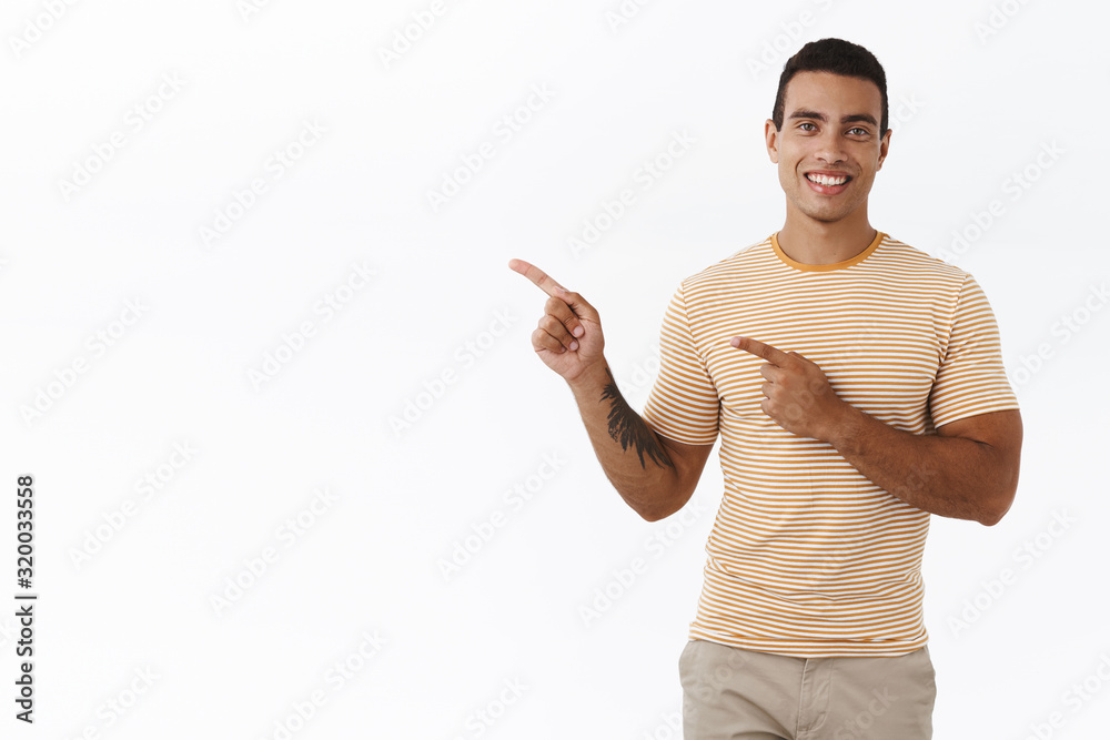 Good-looking modern young man with mascules, tattoo, pointing upper left corner and smiling happily, showing promotion, advertisement banner on white blank space, help with advice Good-looking modern young man with mascules, tattoo, pointing upper left corner and smiling happily, showing promotion, advertisement banner on white blank space, help with advice