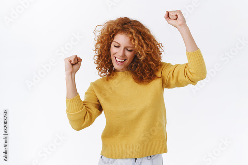 Carefree, attractive modern redhead girl in yellow sweater, dancing as triumphing feeling joy of success, delighted, moving hands, fist pump, close eyes and smiling pleased, white background photo