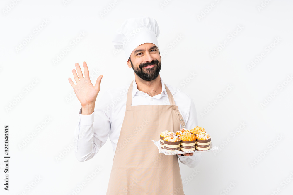 Young man holding muffin cake over isolated white background saluting with hand with happy expression Young man holding muffin cake over isolated white background saluting with hand with happy expression