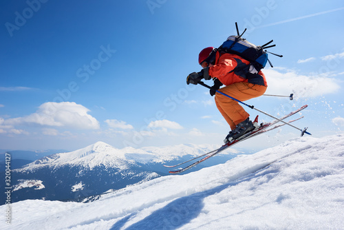 Sportsman skier in skiing equipment jumping in air down steep snowy mountain slope on copy space background of blue sky and highland landscape. Winter risky sports, courage and speed concept.