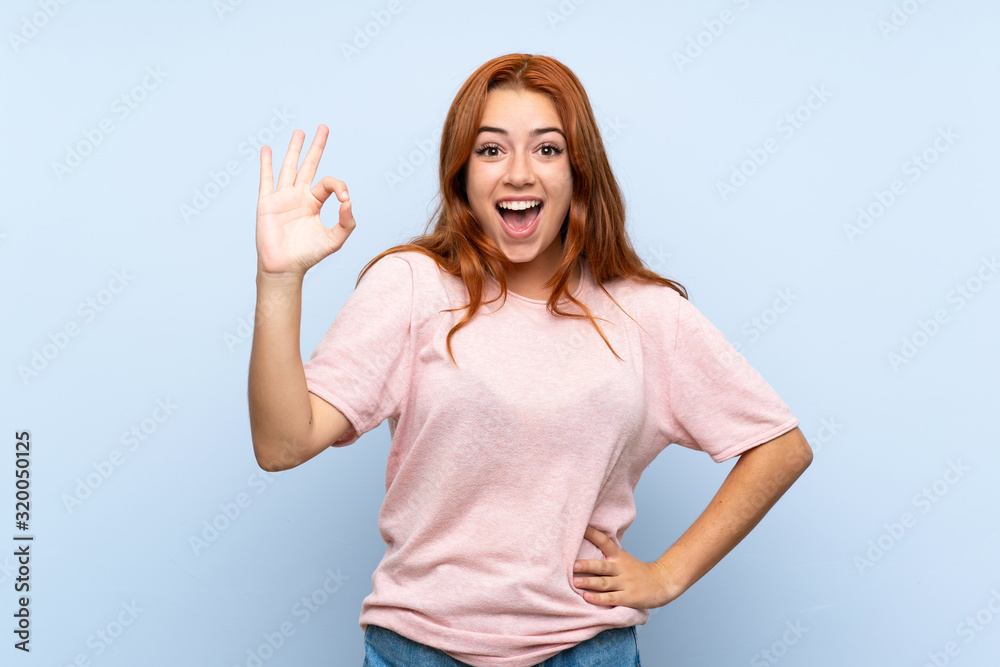 Fototapeta premium Teenager redhead girl over isolated blue background surprised and showing ok sign