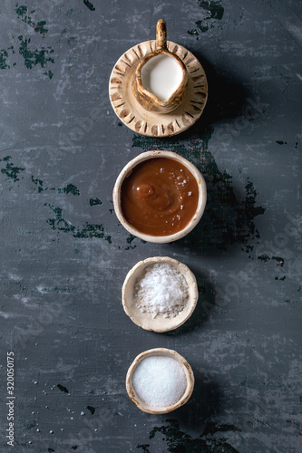Homemade salted caramel sauce with grains of fleur de sel salt. Ingredients in ceramic bowls in row. Grey textured canvas background. Flat lay, space