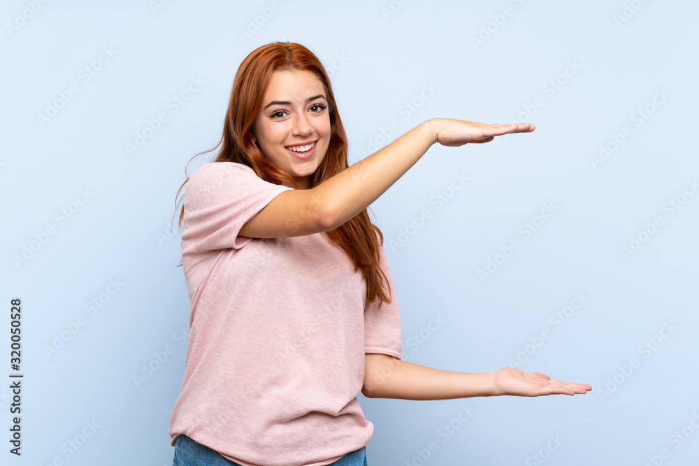 Teenager redhead girl over isolated blue background holding copyspace to insert an ad Teenager redhead girl over isolated blue background holding copyspace to insert an ad