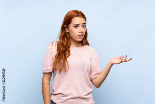 Teenager redhead girl over isolated blue background making doubts gesture
