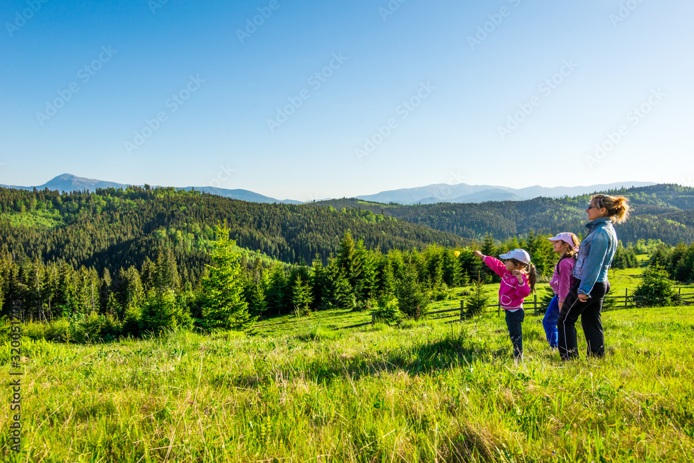 Young mother and two little daughters travelers stand on a slope with a gorgeous view of the hills covered with dense fir forest against the blue sky on sunny warm summer day. Family tourism concept