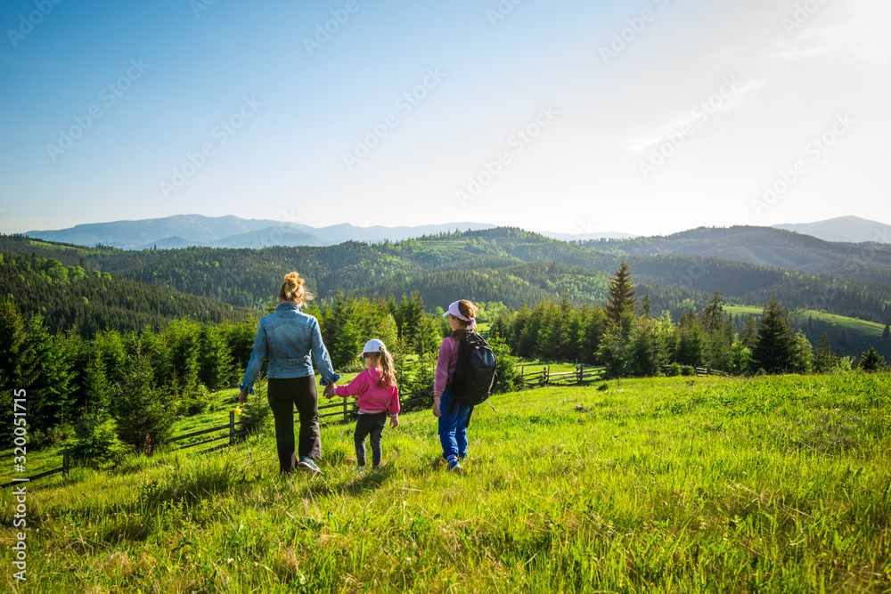 Young mother and two little daughters travelers stand on a slope with a gorgeous view of the hills covered with dense fir forest against the blue sky on sunny warm summer day. Family tourism concept