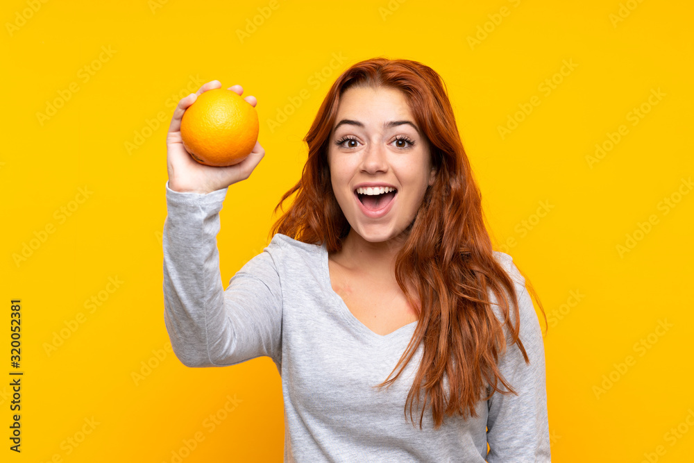 Teenager redhead girl holding an orange over isolated yellow background Teenager redhead girl holding an orange over isolated yellow background