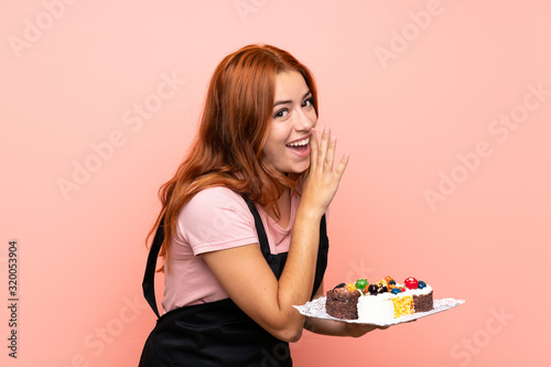 Teenager redhead girl holding lots of different mini cakes over isolated pink background whispering something