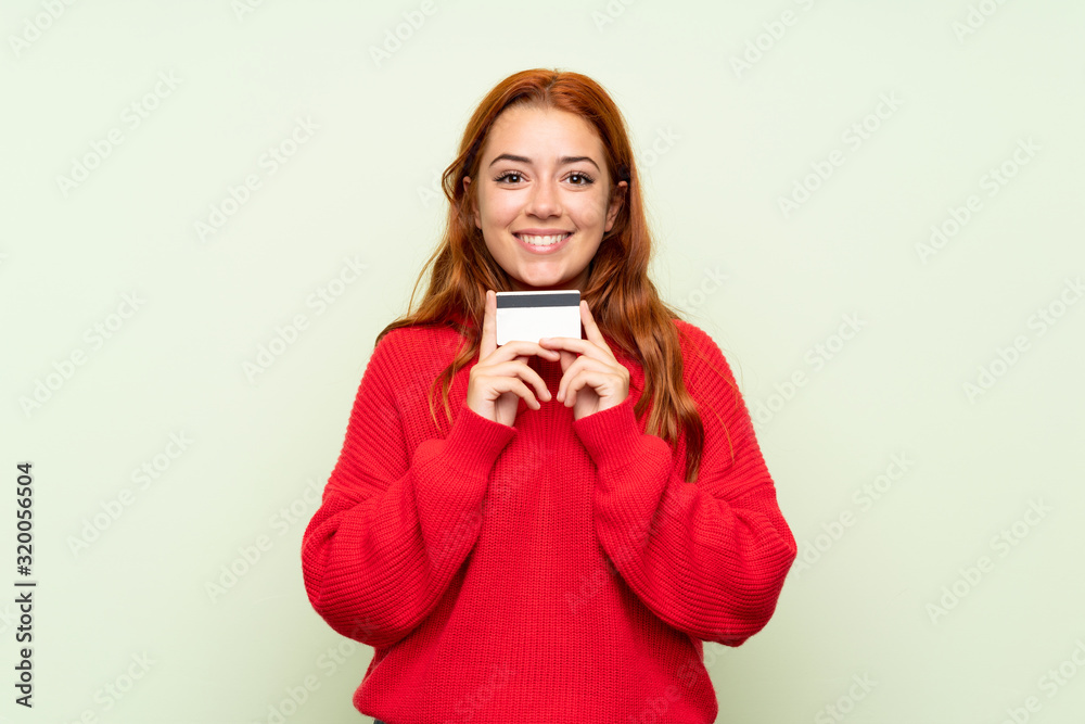 Naklejka premium Teenager redhead girl with sweater over isolated green background holding a credit card