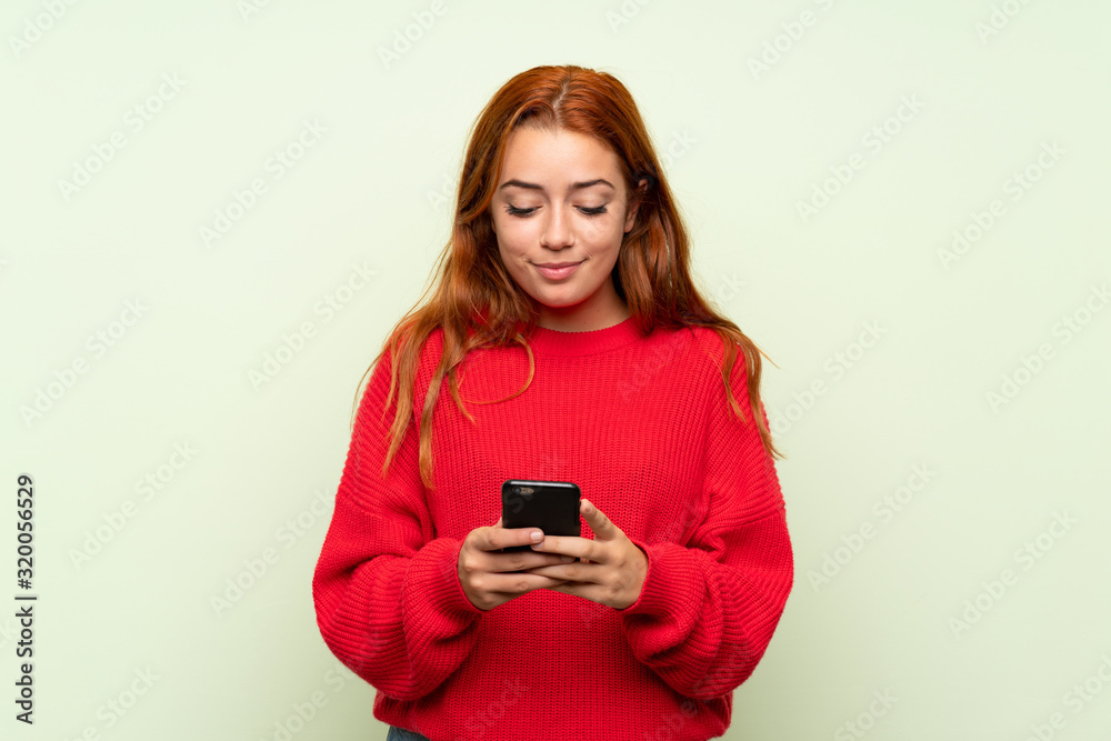 Teenager redhead girl with sweater over isolated green background using mobile phone Teenager redhead girl with sweater over isolated green background using mobile phone