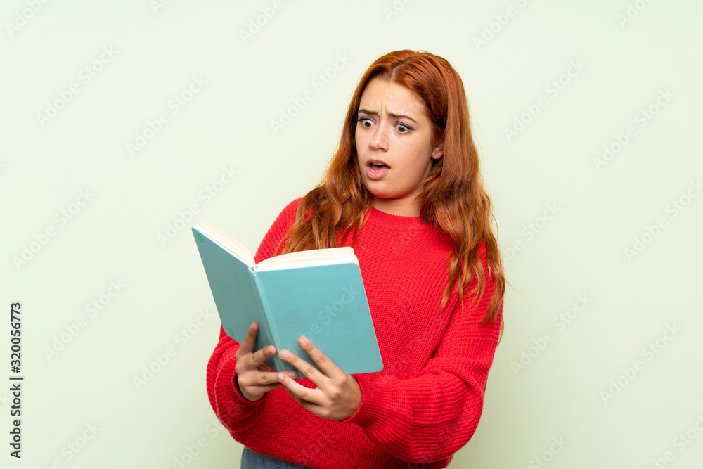 Teenager redhead girl with sweater over isolated green background holding and reading a book Teenager redhead girl with sweater over isolated green background holding and reading a book