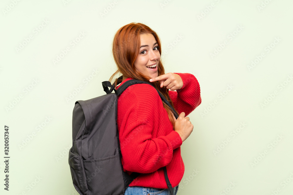 Teenager redhead girl with sweater over isolated green background with backpack Teenager redhead girl with sweater over isolated green background with backpack