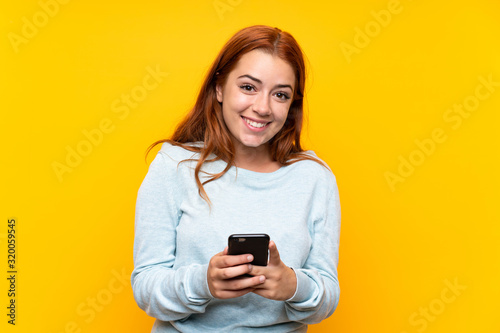 Teenager redhead girl over isolated yellow background sending a message with the mobile © luismolinero