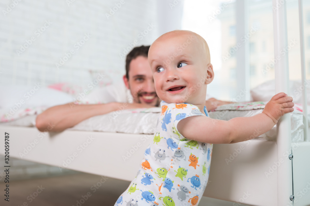 Cute little baby is standing on the edge of the bed holding on to the headboard against the background of his parents. Concept of happy newly made parents. Copyspace Cute little baby is standing on the edge of the bed holding on to the headboard against the background of his parents. Concept of happy newly made parents. Copyspace