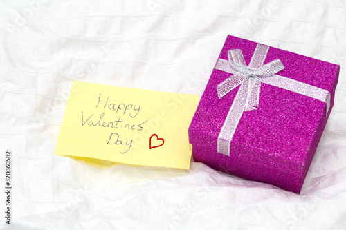 closeup of a little paper note with the text happy valentines day written in it, on the white sheets of a bed with a present box